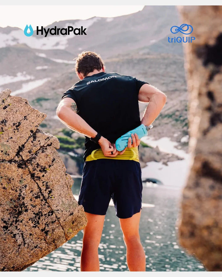 A man stands outdoors in a rocky mountain setting, viewed from behind, wearing a black Salomon running shirt and dark shorts while adjusting a light blue hydration pack. The Salomon brand name is visible on the back of his shirt, and the HydraPak logo appears in the top left corner.