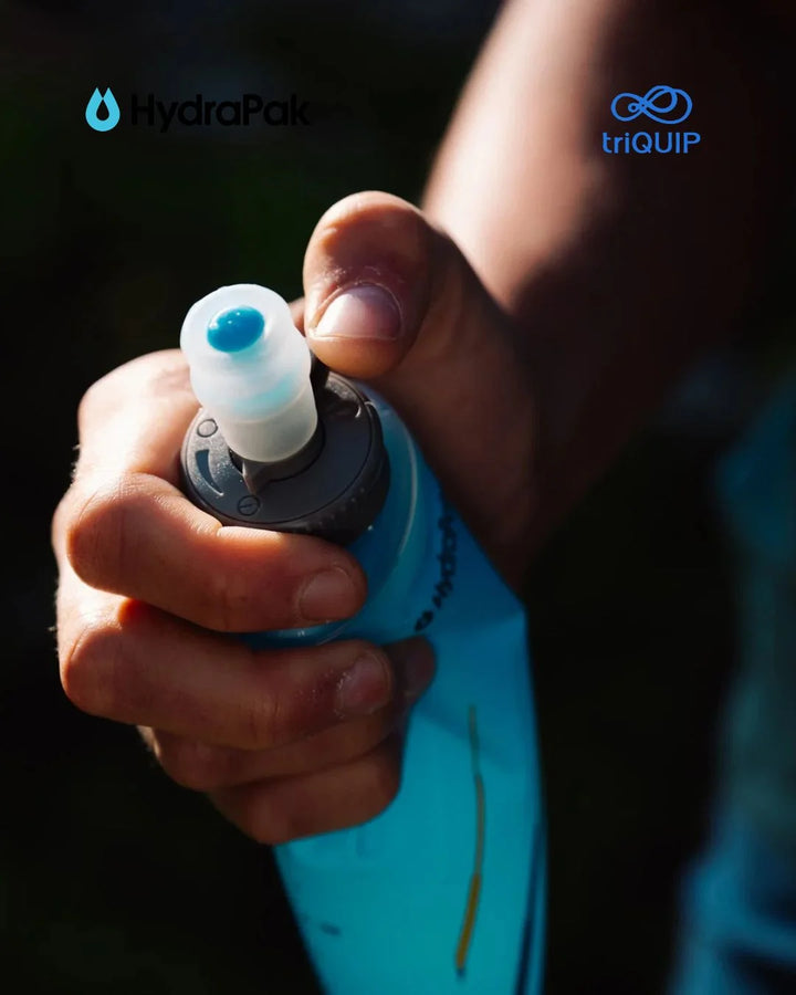 A close-up of a person's hand holding a light blue HydraPak hydration tube with a flip-top cap, with the HydraPak and triQuIP logos visible in the top corners.