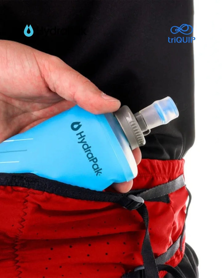 A close-up of a person's hand holding a bright blue Hydrapak water bottle with a gray and clear straw, shown tucked into a red hydration pack with black straps.
