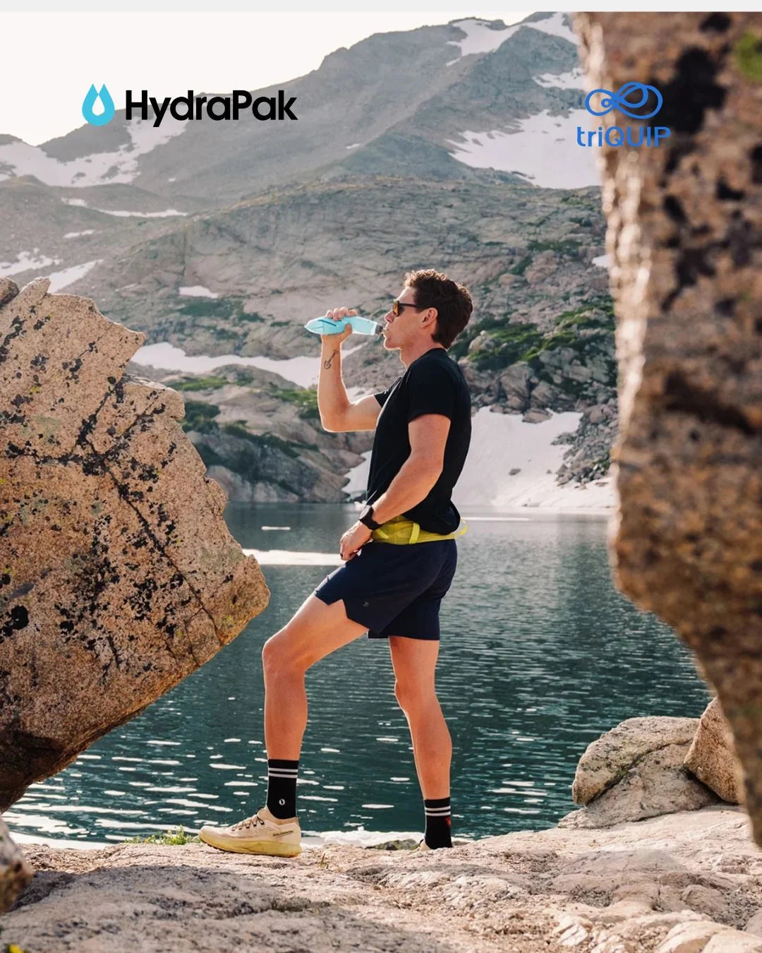 A man in athletic wear stands by a mountain lake drinking from a blue water bottle, with the HydraPak logo in the top left and the tri-UP logo in the top right.