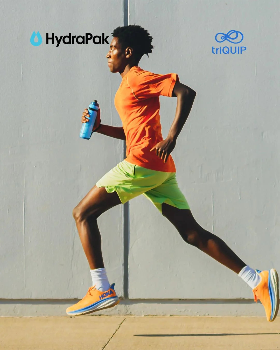 A man running in profile wearing an orange t-shirt, neon green shorts, and orange running shoes, holding a blue water bottle. The background is a plain light gray wall.