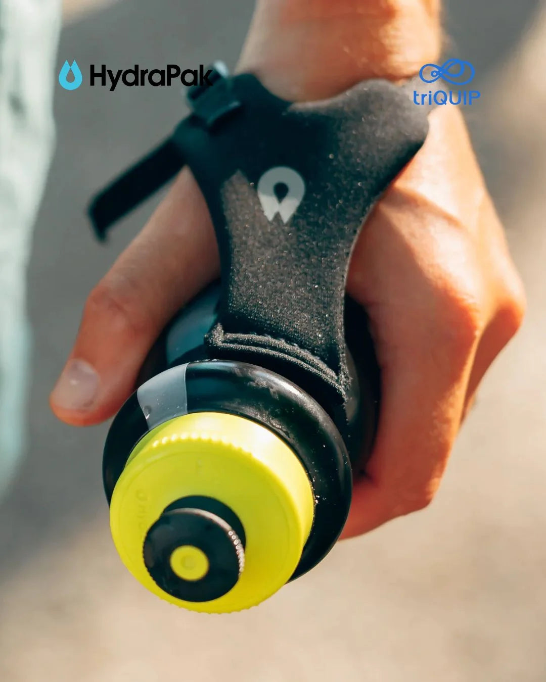 A close-up of a hand holding a HydraPak water bottle with a black ergonomic handle and a bright yellow cap, outdoors with a blurred background.