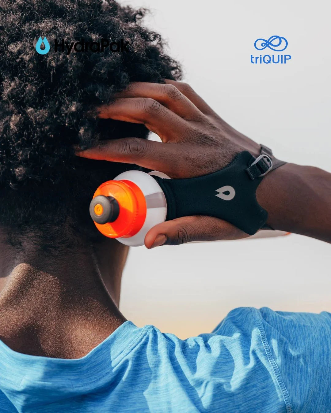 A close-up lifestyle shot of a person outdoors wearing a black wrist strap device that holds a white and orange water bottle. The person has curly dark hair and is wearing a blue shirt, with the YardPak and triQUIP logos visible in the top corners.