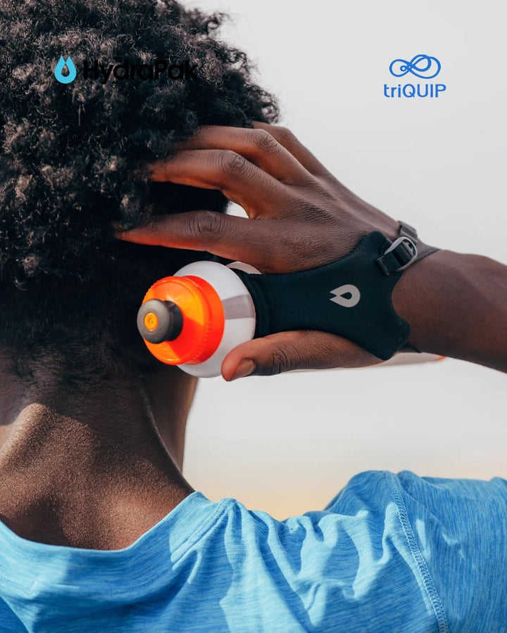 A close-up lifestyle shot of a person outdoors wearing a black wrist strap device that holds a white and orange water bottle. The person has curly dark hair and is wearing a blue shirt, with the YardPak and triQUIP logos visible in the top corners.