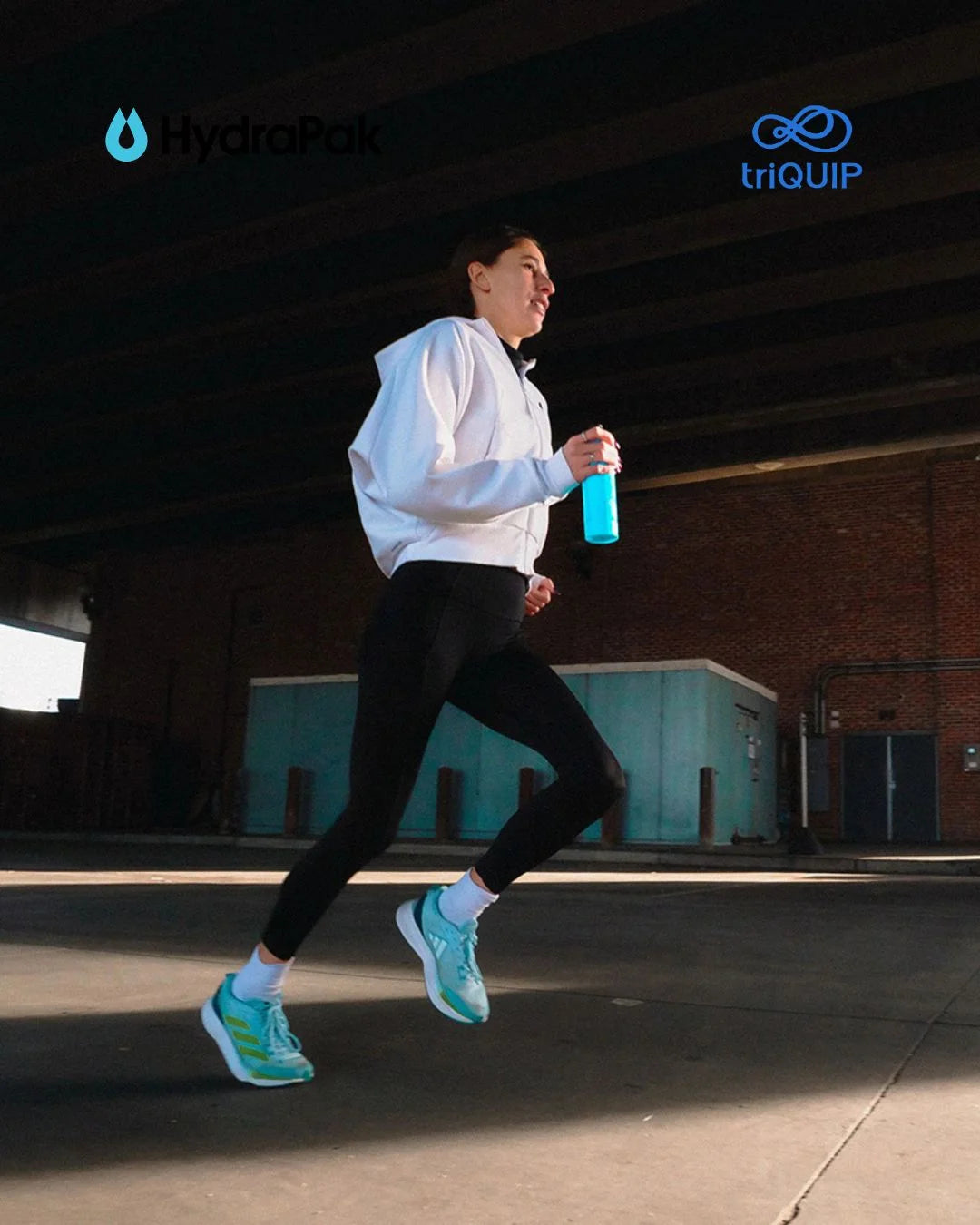 A man running outdoors under a brick structure wearing a white hoodie, black leggings, and bright blue running shoes, while holding a blue HydroPak water bottle. The HydroPak logo is visible in the top left, and the triQUIP logo is visible in the top right.