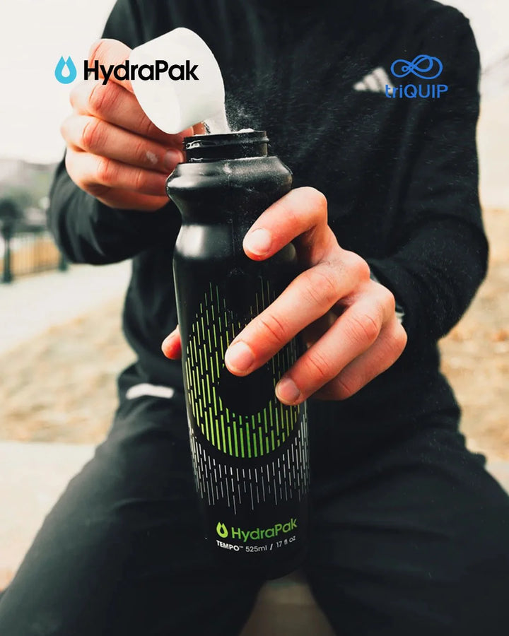 A person in black athletic clothing holds a black HydraPak insulated water bottle while pouring liquid from a white scoop into it. The HydraPak logo appears on the bottle, and a HydraPak and Equip watermark is visible in the upper corners.