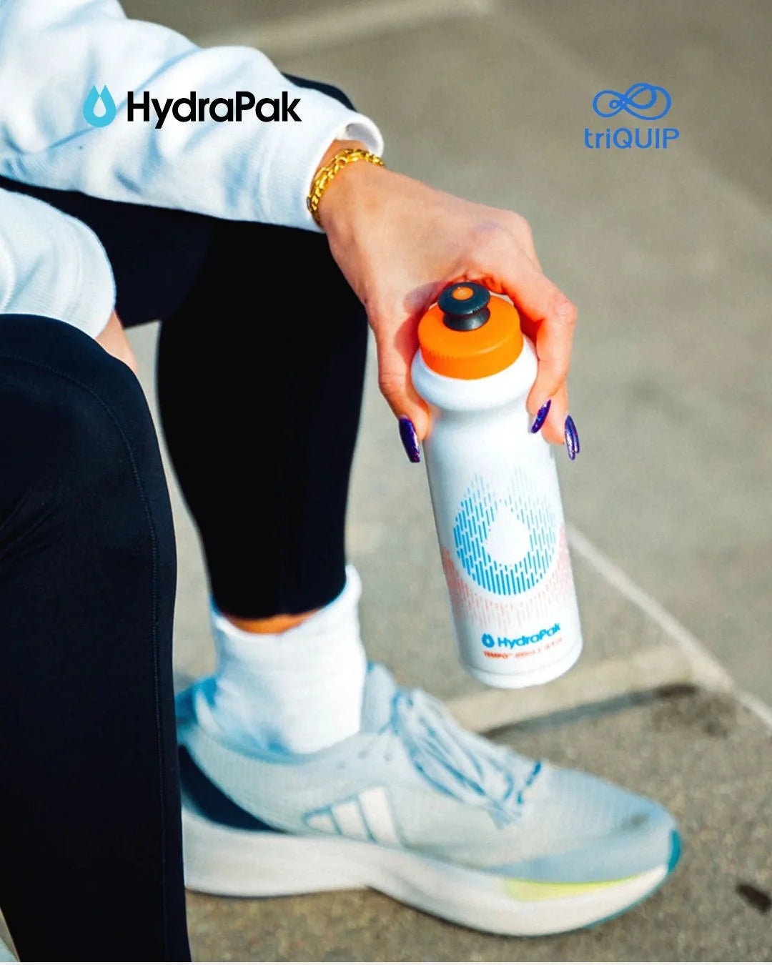 A person in athletic wear holds a white HydraPak water bottle with an orange cap. The scene includes visible HydraPak and triQUIP branding, with the person seated outdoors on a concrete surface.