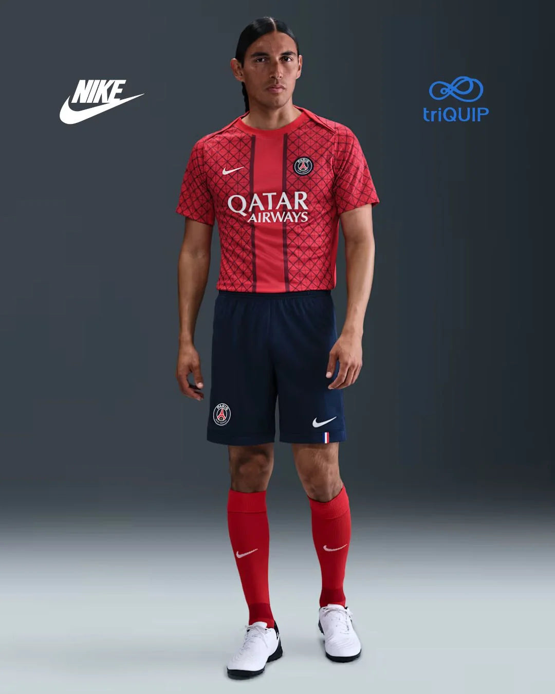 A male model wearing a red and navy Paris Saint-Germain soccer kit by Nike, featuring a patterned red jersey with Qatar Airways sponsor, navy shorts, red socks, and white Nike cleats, set against a dark gray studio background with Nike and triQUIP logos.
