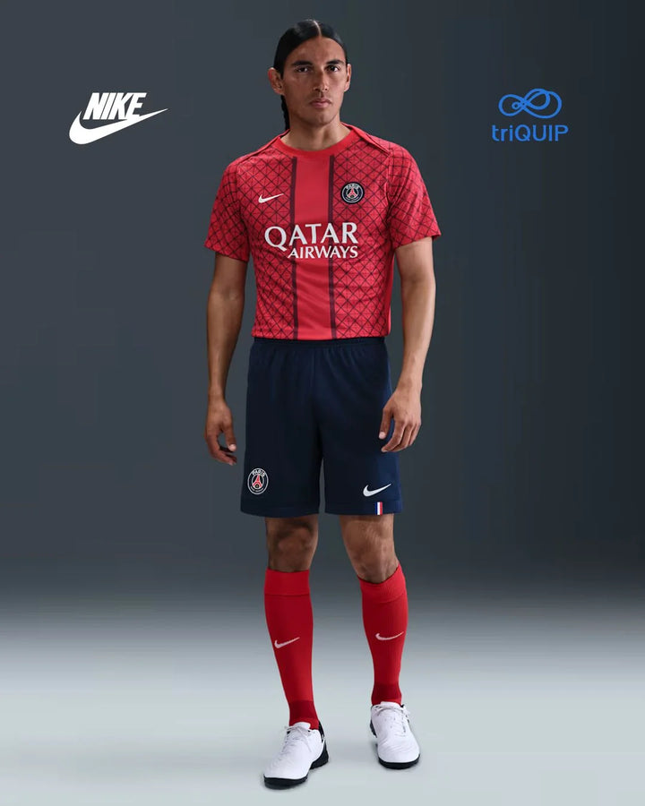 A male model wearing a red and navy Paris Saint-Germain soccer kit by Nike, featuring a patterned red jersey with Qatar Airways sponsor, navy shorts, red socks, and white Nike cleats, set against a dark gray studio background with Nike and triQUIP logos.