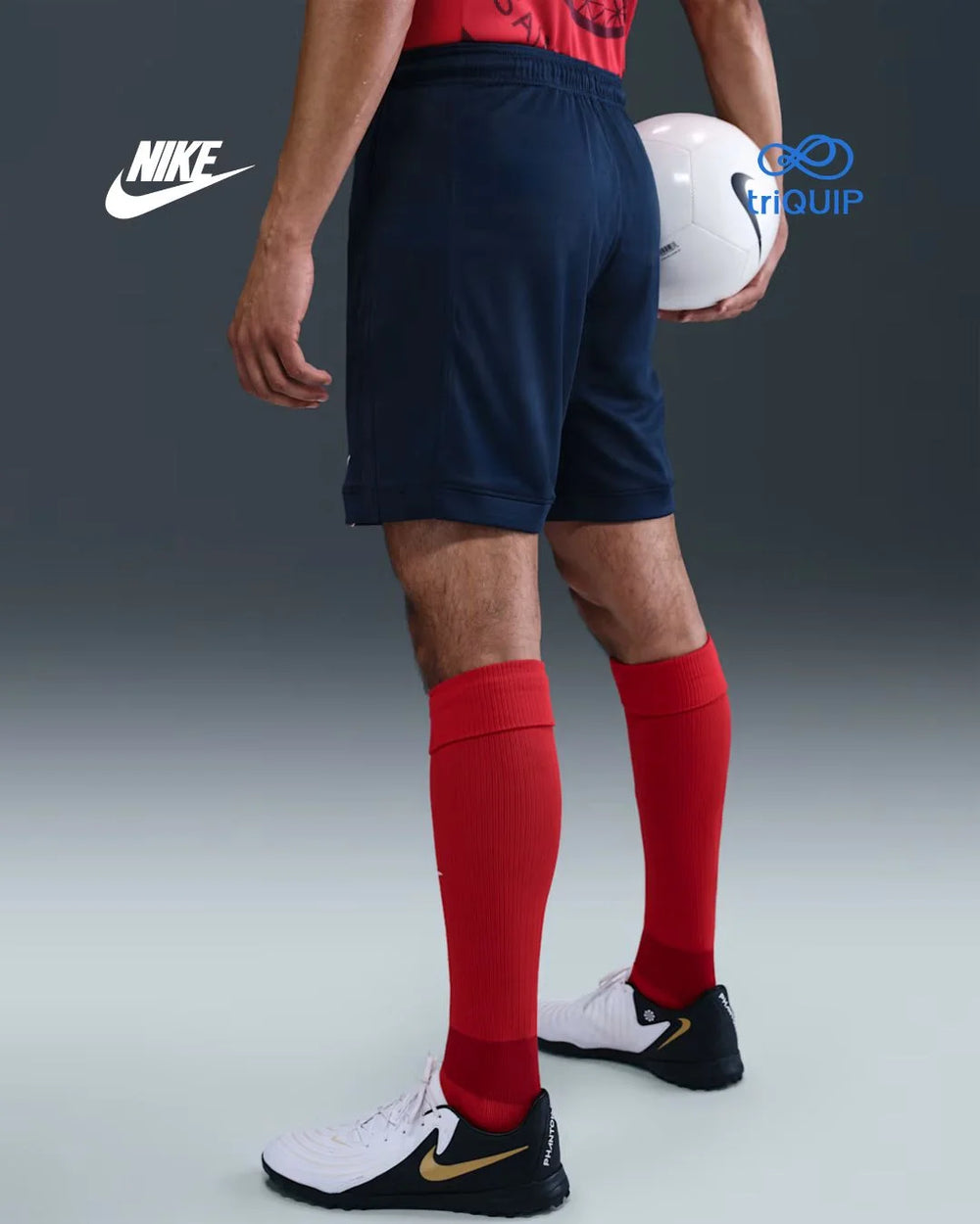 A model from the waist down wearing navy blue Nike soccer shorts, bright red knee-high socks, and white soccer cleats, holding a white soccer ball. The Nike logo is visible on the shorts and shoes, and a Trioup logo is in the upper right corner.
