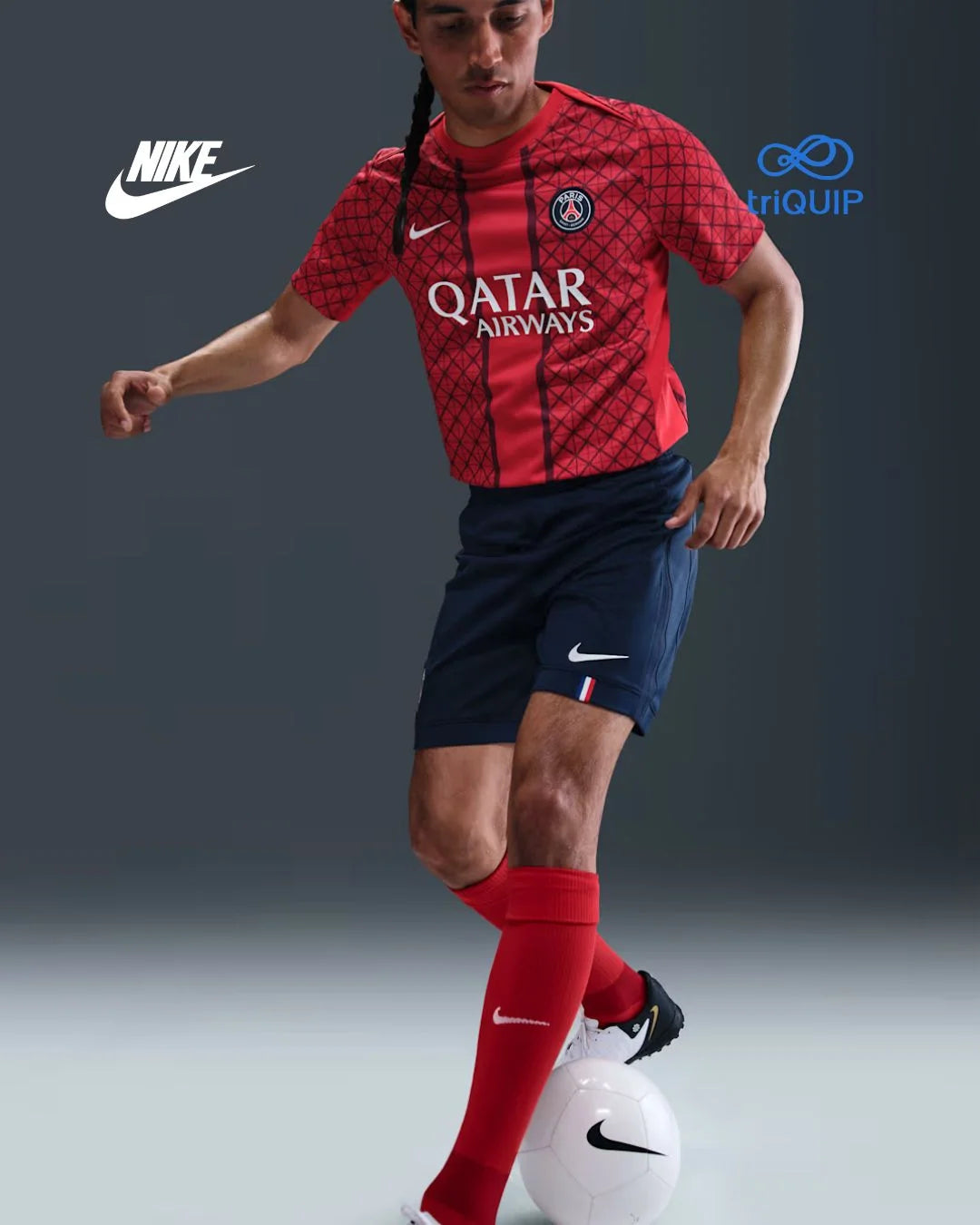 A male soccer player in a red Paris Saint-Germain jersey with 'Qatar Airways' sponsor, navy shorts, and red Nike socks, posed with a soccer ball against a dark gray studio background. Nike and Triquip logos are visible in the upper corners.